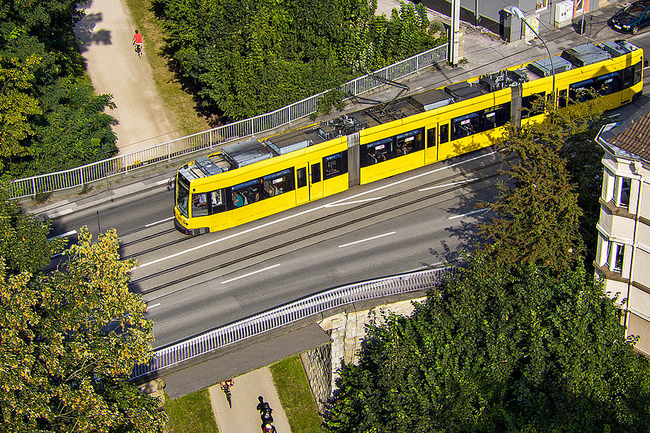 Fotomontage einer gelben Straßenbahn, die über eine Brücke in Essen fährt. Unterhalb der Brücke sind Fahrradfahrer zu sehen.