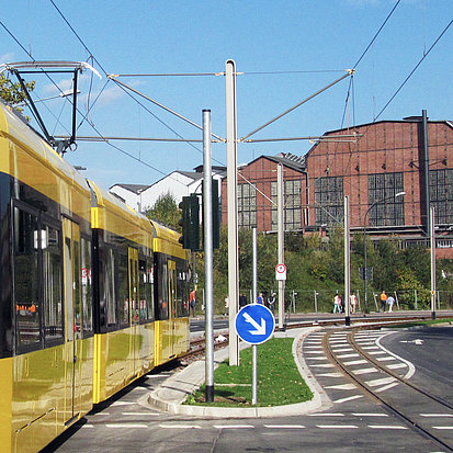 Beispielbild eines Standardmastes für Straßenbahnen, der auf einer Gründfläche in der Mitte einer Straße steht. Links fährt eine gelbe Straßenbahn vorbei.